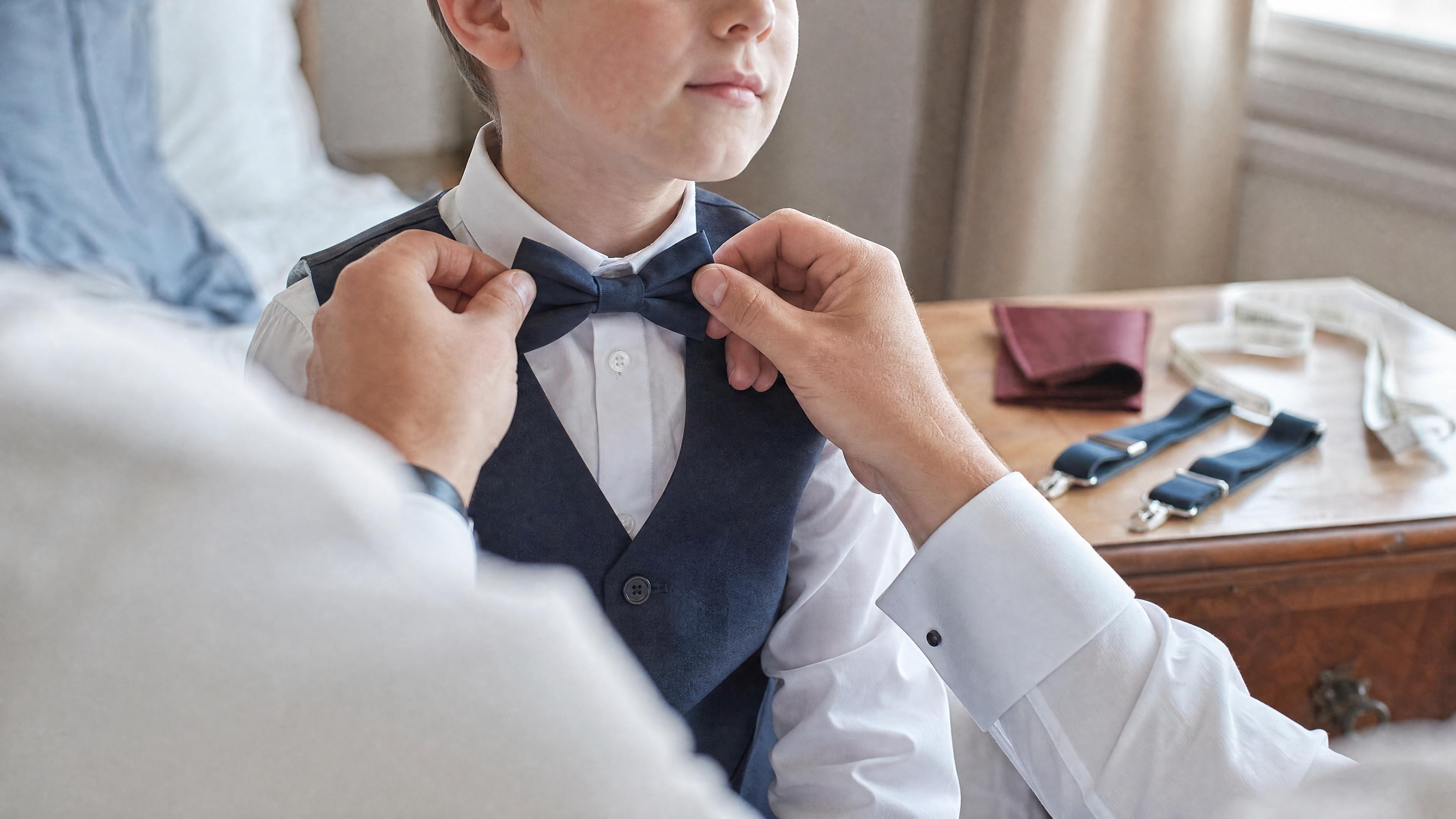 Un adulte ajuste un nœud papillon sur un enfant en chemise blanche, avec un mètre ruban visible sur une commode, dans une chambre lumineuse avant une cérémonie Imagen